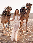 A young woman wearing a long white wedding dress with two camels