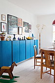 Candles and ornaments on blue sideboard below pictures on wall, wooden rocking horse, dining table and chairs in foreground