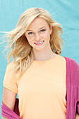 A young blonde woman wearing an apricot t-shirt and a purple jumper against a blue background