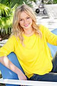 A young blonde woman wearing a yellow jumper sitting outside on a upholstered bench
