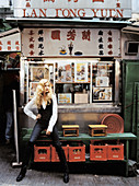 A blonde woman wearing a white shirt, a black waistcoat and black trousers standing outside a Chinese take-away