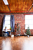 Frau am Tisch vor Fenster, daneben Leiter mit Weihnachtsdekoration, Rehfigur und kleiner Tannenbaum vor Backsteinwand