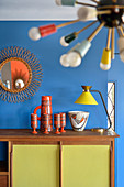 Collection of ceramics and table lamp on retro sideboard below sunburst mirror on blue wall