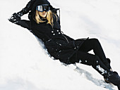 A blonde woman wearing ski goggles, a black winter coat, leggings and boots in the snow