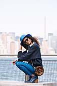 A dark-haired woman wearing a hat, sunglasses, jumper and jeans