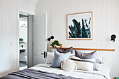 Scatter cushions on double bed against narrow shelf in bedroom with white wood-clad walls