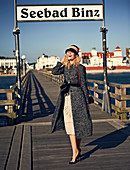 Junge Frau mit Hut im Mantel aus Wollmix und Midikleid vor dem Seebad Binz