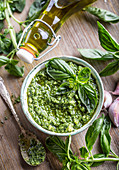 Pesto in einem Schälchen, frisches Basilikum und Olivenöl auf Holztisch