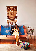 Young woman with dog sits on sofa with blanket, ethnic wall hanging above