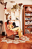 Young woman sits with dog in armchair in front of fireplace shelf
