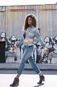 A dark-haired woman wearing denim dungarees and a denim jacket standing against a graffitied wall