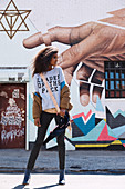 A dark-haired woman wearing a t-shirt, jeans and a jacket standing against a wall painted with street art
