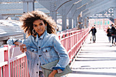 A dark-haired woman wearing jeans and a denim jacket standing on a bridge