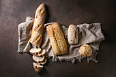 Variety of loafs fresh baked artisan white and whole grain bread on linen cloth over dark brown texture background