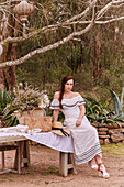 A brunette woman wearing a white, embroidered, off-the-shoulder maxi dress sitting at a garden table