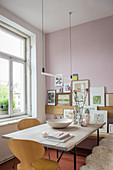 A pendant lamp above a wooden dining table with a picture gallery in the background on a pink wall