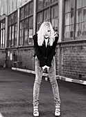 A blonde woman wearing a black jacket and jeans in front of an industrial building (black-and-white shot)