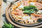 Freshly baked pizza with artichokes, smoked turkey ham, olives, cream cheese and basil over rustic wooden background