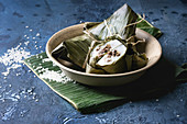 Asian rice piramidal steamed dumplings from rice tapioca flour with meat filling in banana leaves served in ceramic bowlwith rice above over blue texture background
