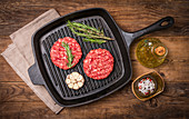 Raw burgers from organic beef with garlic and rosemary in a frying pan on an old wooden table, top view