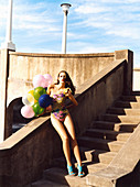 A brunette woman on a flight of steps wearing a bathing suit and holding balloons