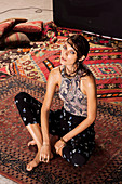 A young woman wearing a Paisley patterned summer blouse and black floral patterned trousers sitting on a rug