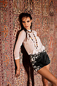 A young woman wearing a white blouse and black shorts standing against a hanging rug