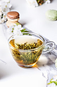 Grüner Tee mit Teeblume in Glastasse, dahinter Macarons und Blüten