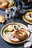 Variety of homemade bagels with sesame seeds, cream cheese, pesto sauce, eggs, radish, herbs served on ceramic plate