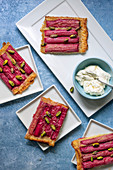 Servings of rhubarb tart sprinkled with pistachio nuts on dessert plates with a bowl of cream