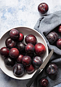 A bowl of ripe red plums and a knife with scattered plums on a blue napkin