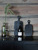 Black chopping boards, bottle of wine and glasses against wooden wall