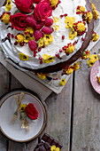 Chocolate cake with eatable flowers yellow and red roses, raspberries and whipped cream on a wooden table with plates