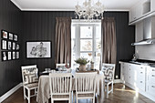 Dining table with linen tablecloth and chairs below chandelier in kitchen with dark walls