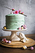Easter cake decorated with pastel mini eggs and cheery blossom on a white bunny rabbit cake stand