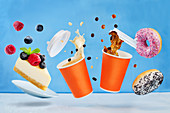 Fliegende Kaffeebecher, Donuts und Käsekuchen vor blauem Hintergrund