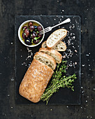 Italian ciabatta bread cut in slices on wooden chopping board with herbs, garlic and olives