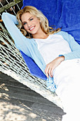 A blonde woman wearing a white top and a light blue cardigan lying in a hammock