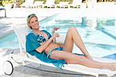 A blonde woman wearing a turquoise patterned bikini on a lounger by a pool