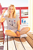 A blonde woman wearing a striped top and shorts sitting against a beach hut