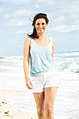 A brunette woman on a beach wearing shorts and a vest top