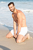 A young topless man kneeling in the sand wearing shorts