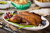A platter of roast duck with a holiday meal in the background on a warm, rustic wood tabletop