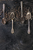 Variety of different black peppers allspice, pimento, monks pepper, peppercorns in vintage spoons over old black iron texture background