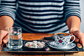 A woman serving Turkish coffee in a traditional Turkish coffee cup on a traditional copper tray, a glass of water with a rose petal inside and Turkish delights accompany