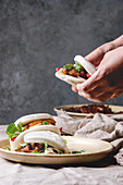 Man's hands hold asian sandwich steamed gua bao buns with pork belly, greens and vegetables served in ceramic plate