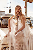 A blonde woman wearing a white pleated dress sitting on the bonnet of a car