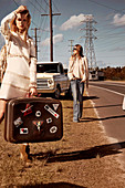 Two women with a suitcase at the edge of a road with a car parked in the background