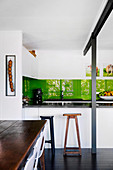 Fitted kitchen with green back wall, bar stool at the counter, rustic dining table in the foreground