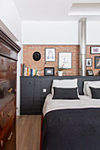 Doppelbett vor schwarzem Sideboard an Ziegelwand im Schlafzimmer, im Vordergrund Antik Kommode